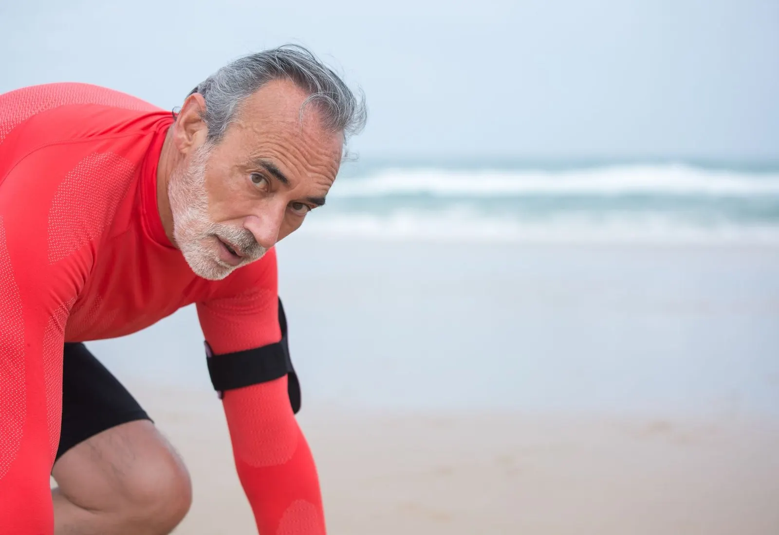 Homme d’une soixantaine d’années en tenue de sport, accroupi sur une plage face à la mer