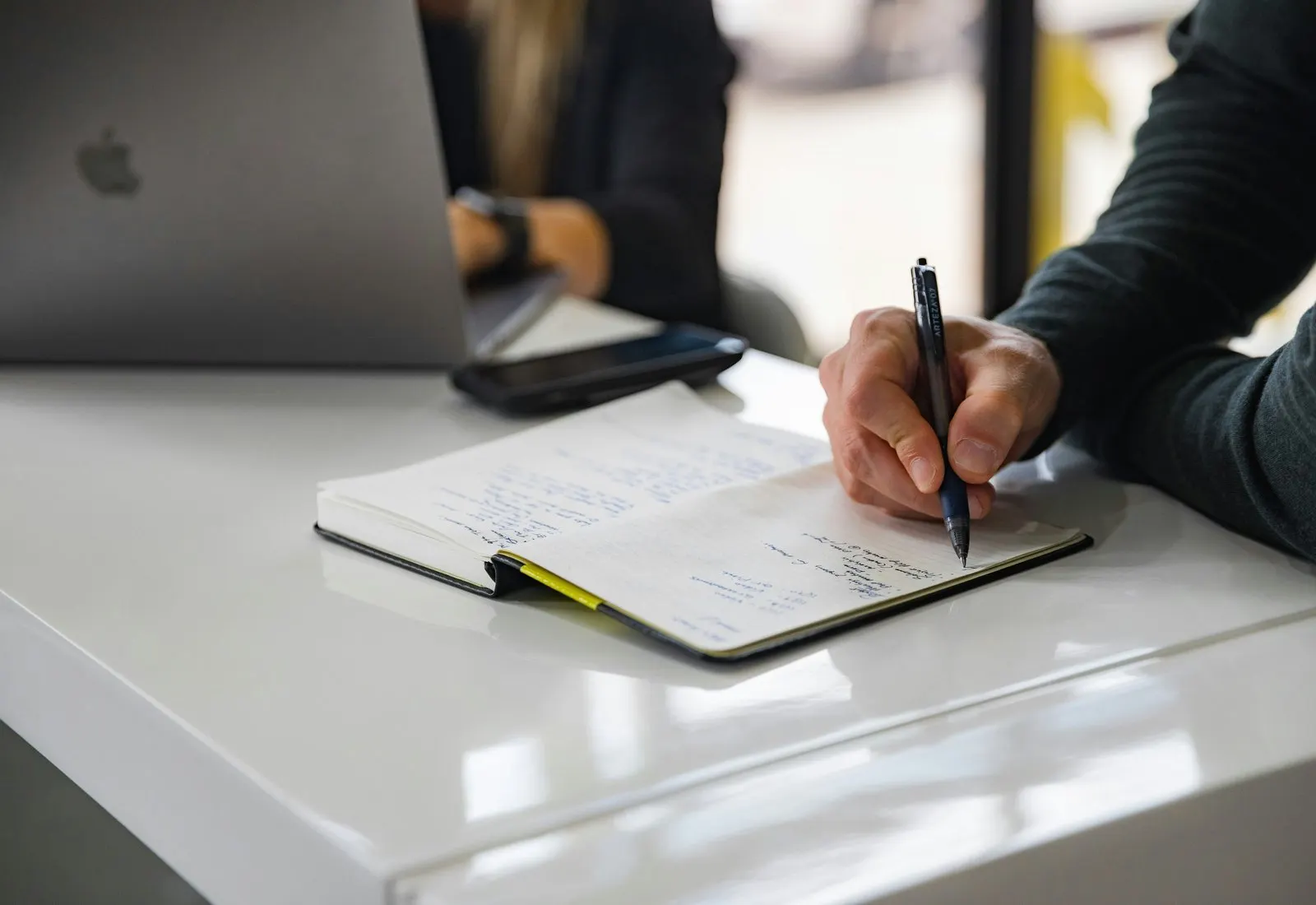 Main qui prend des notes dans un carnet, à côté d’un ordinateur portable