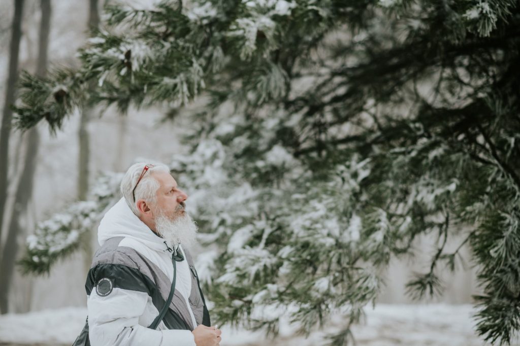 Homme senior à barbe blanche marchant en veste d'hiver dans une forêt enneigée de pins