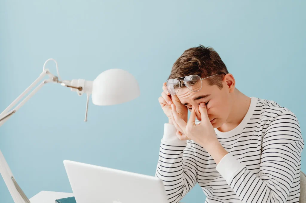 Un jeune homme se frotte les yeux devant son ordinateur portable, lampe de bureau allumée